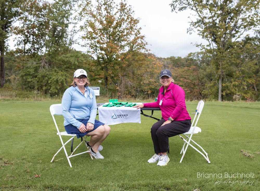 Two women sit at a table on a golf course during an outdoor event on a cloudy autumn day.