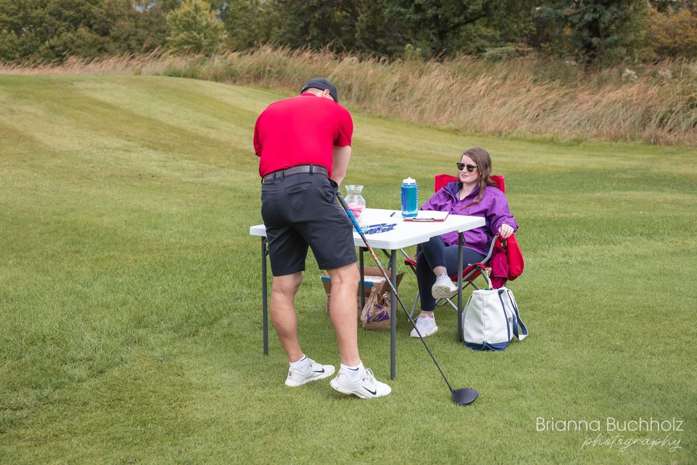 Two golfers pause on a green fairway, sharing a moment at a small table with drinks during a casual golf outing.