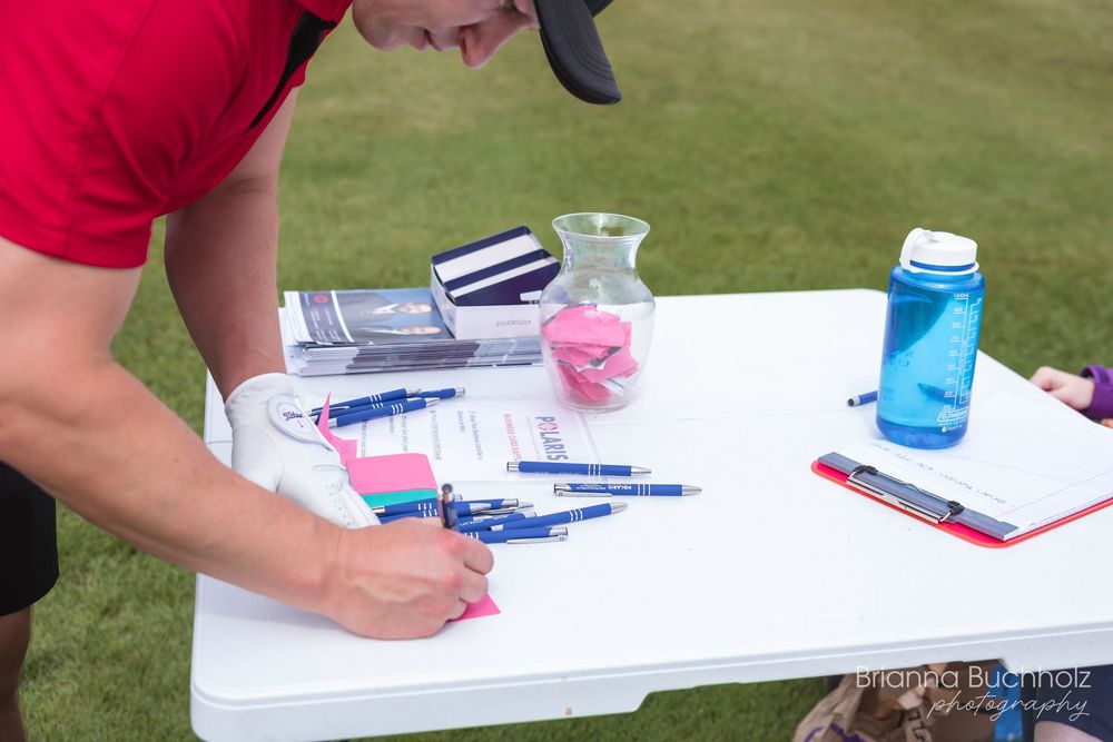 Person in red shirt writing at outdoor white table with pens, notebooks, and a blue water bottle on green grass.