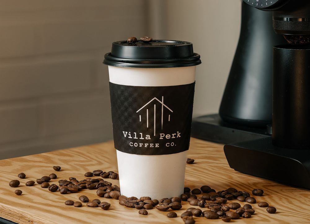 Villa Perk Coffee Co. branded paper cup surrounded by coffee beans on a wooden surface.