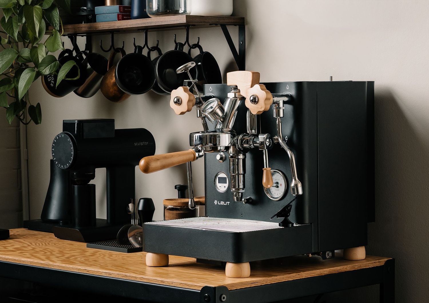 Black espresso machine with wood accents on a wooden counter, surrounded by hanging pots and a coffee grinder.
