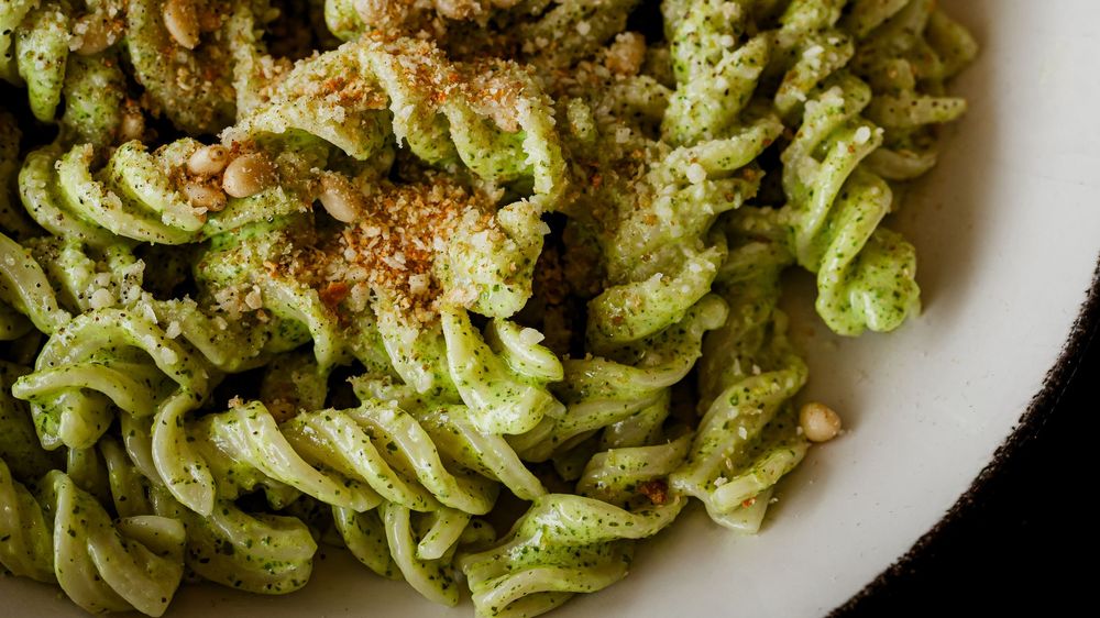 Close-up of fusilli pasta coated in vibrant green pesto sauce, topped with pine nuts and breadcrumbs in a white bowl.