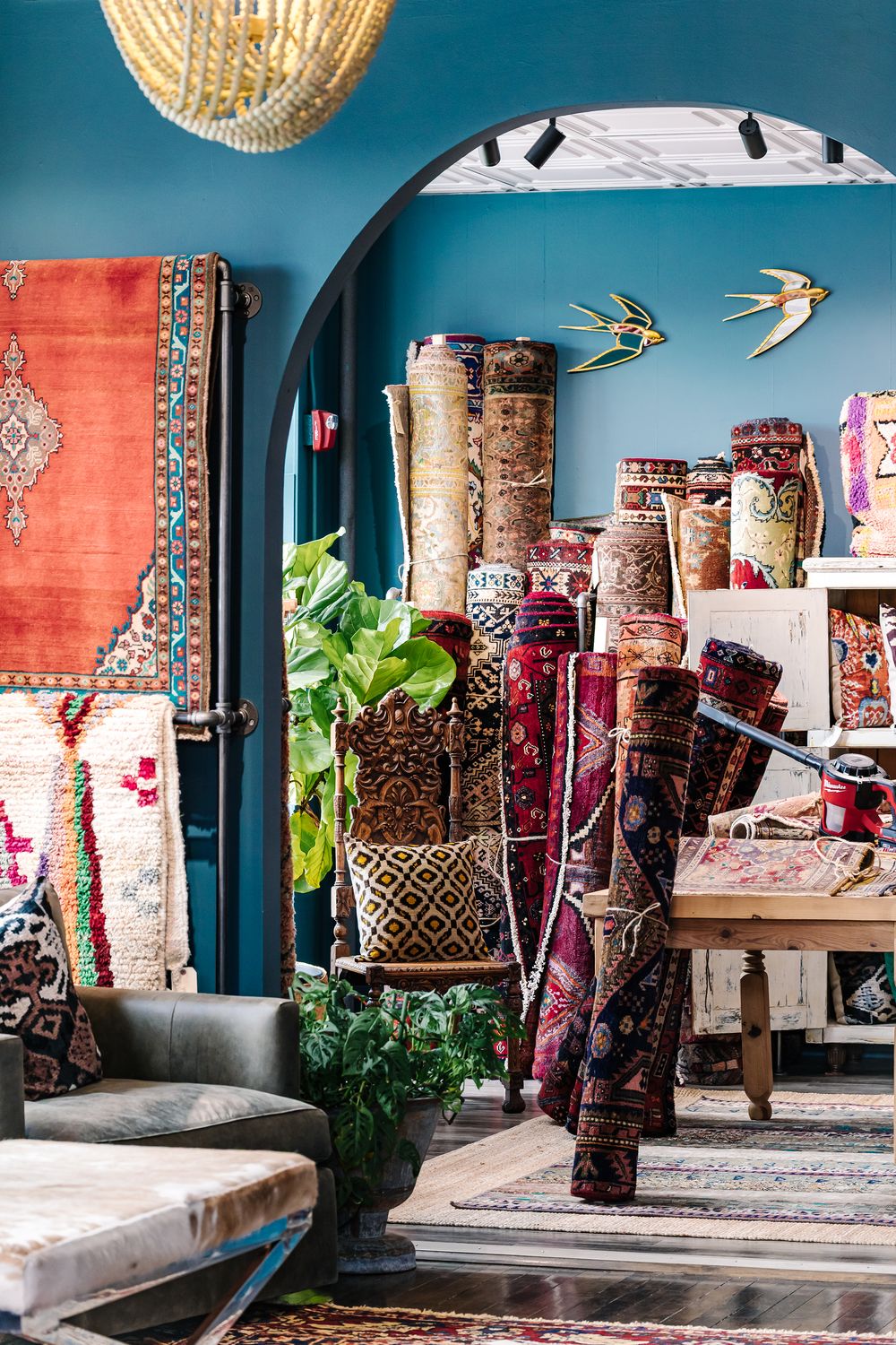 Colorful rolled rugs displayed in a bohemian rug shop with teal walls, arched doorway, and eclectic decor.