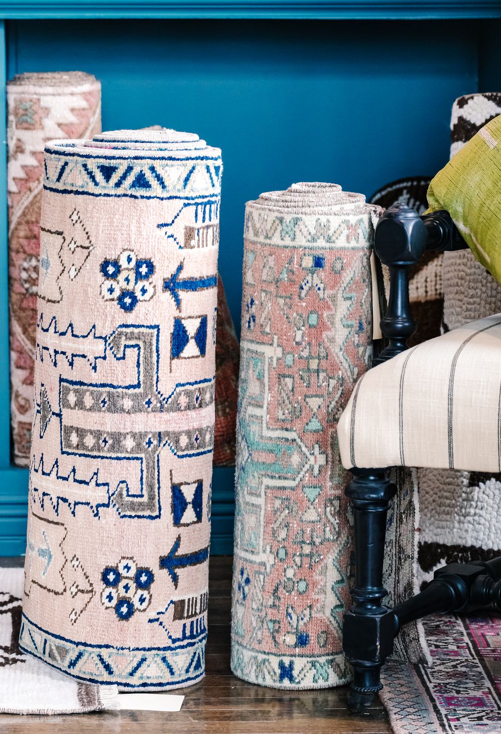 Rolled decorative rugs with boho geometric patterns in pink, blue, and mint displayed against a teal wall.
