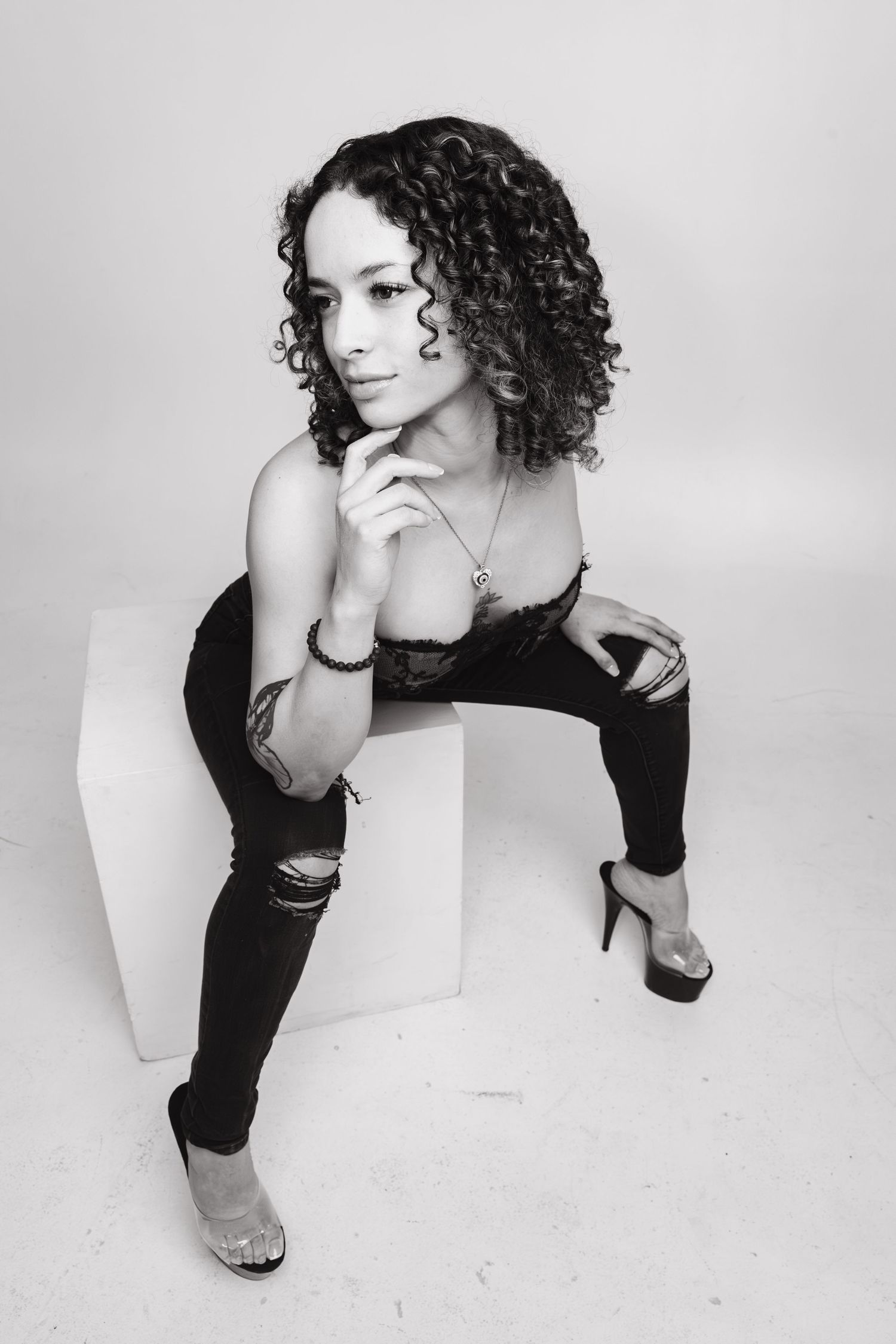 Black and white portrait of a curly-haired woman in ripped jeans and heels posing on a white cube.
