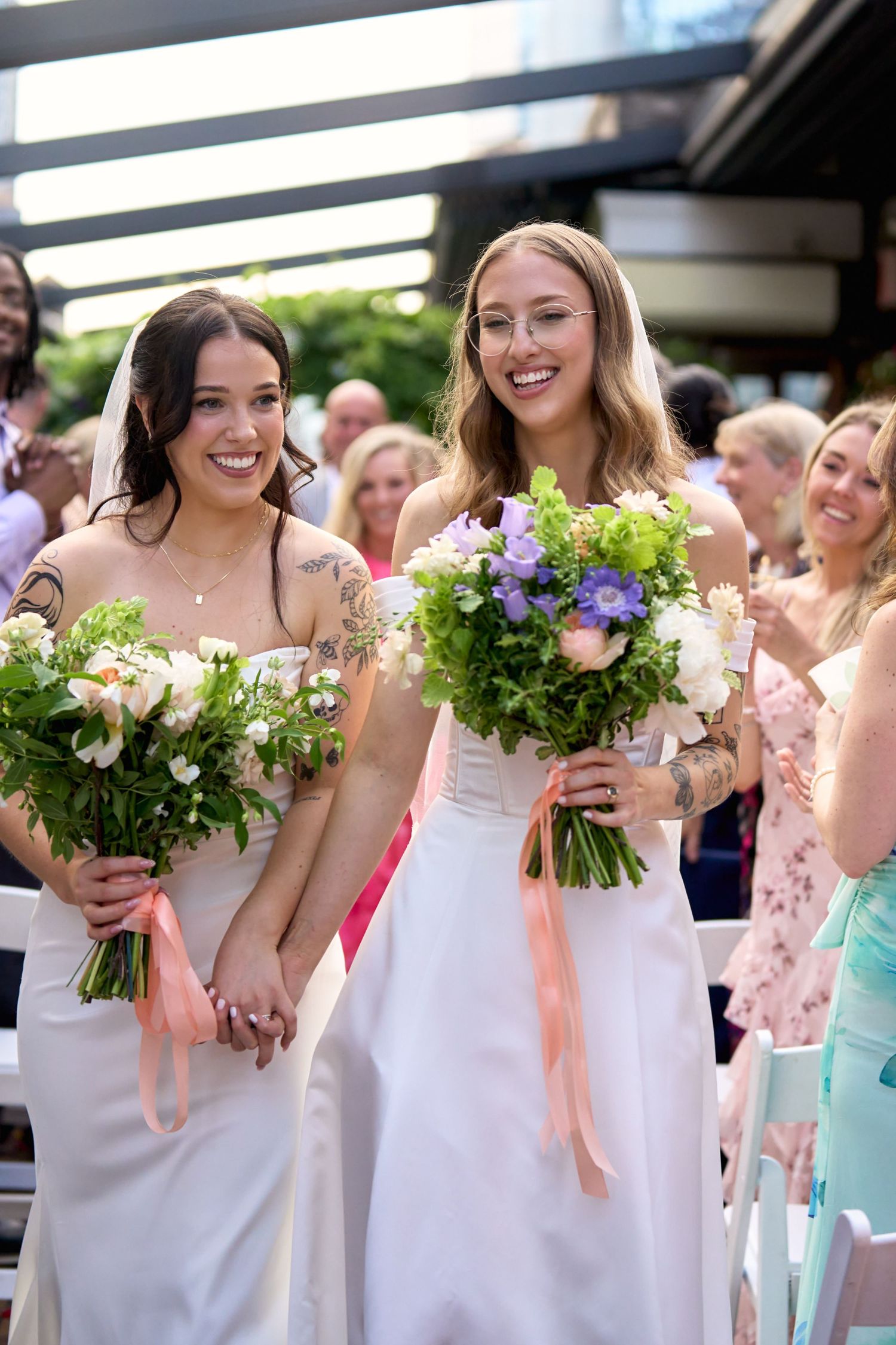 Two brides in white gowns holding floral bouquets walk hand in hand down the aisle at their outdoor wedding ceremony.