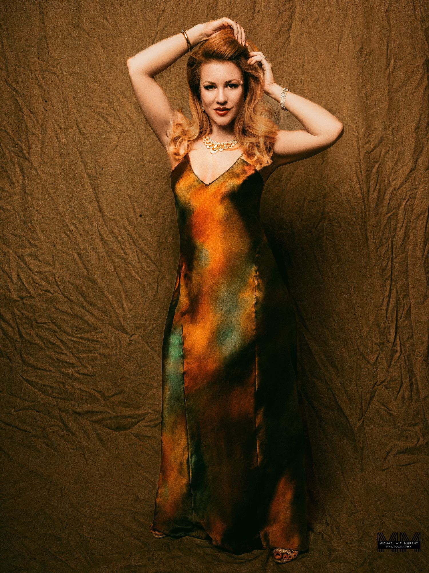 Woman in flowing orange and green tie-dye gown poses elegantly against a textured brown backdrop.