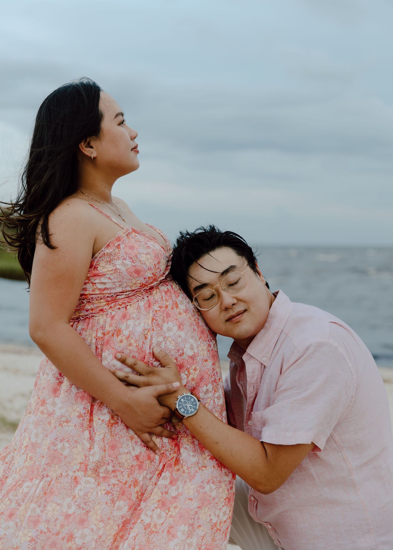 A man kneels at a pregnant woman's belly on a beach, both dressed in pink, sharing a tender maternity moment by the sea.