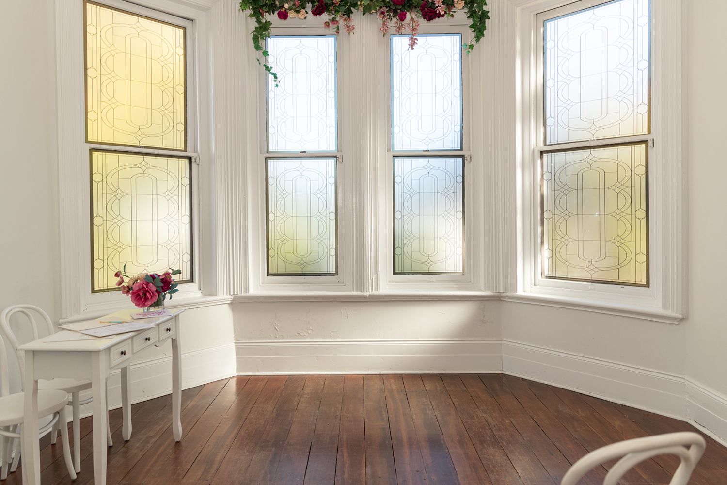 Bright white room with hardwood floors, three frosted windows, a white desk with flowers, and holiday garland above.