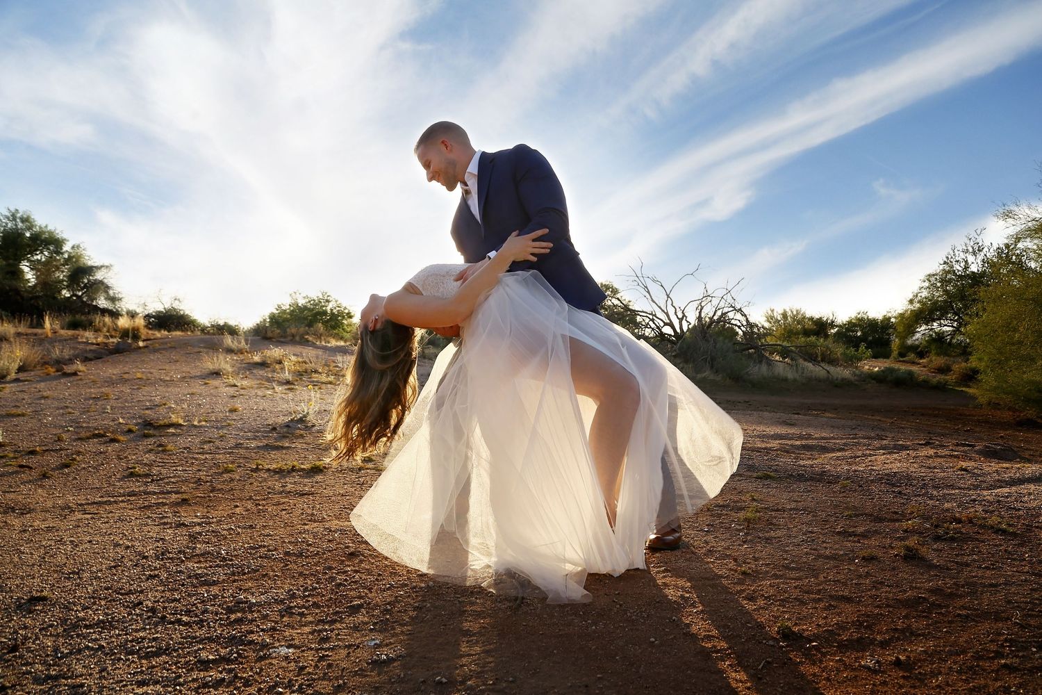 Groom dips his bride in a flowing white gown on a desert trail beneath a bright blue sky with wispy clouds.
