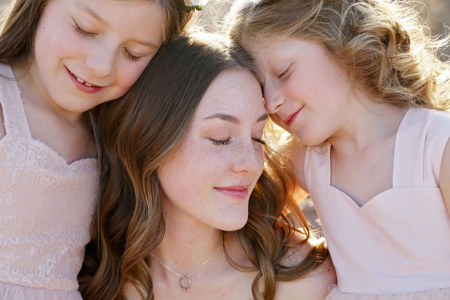 Two girls in blush pink dresses rest their heads against a young woman with wavy hair in warm, golden sunlight.
