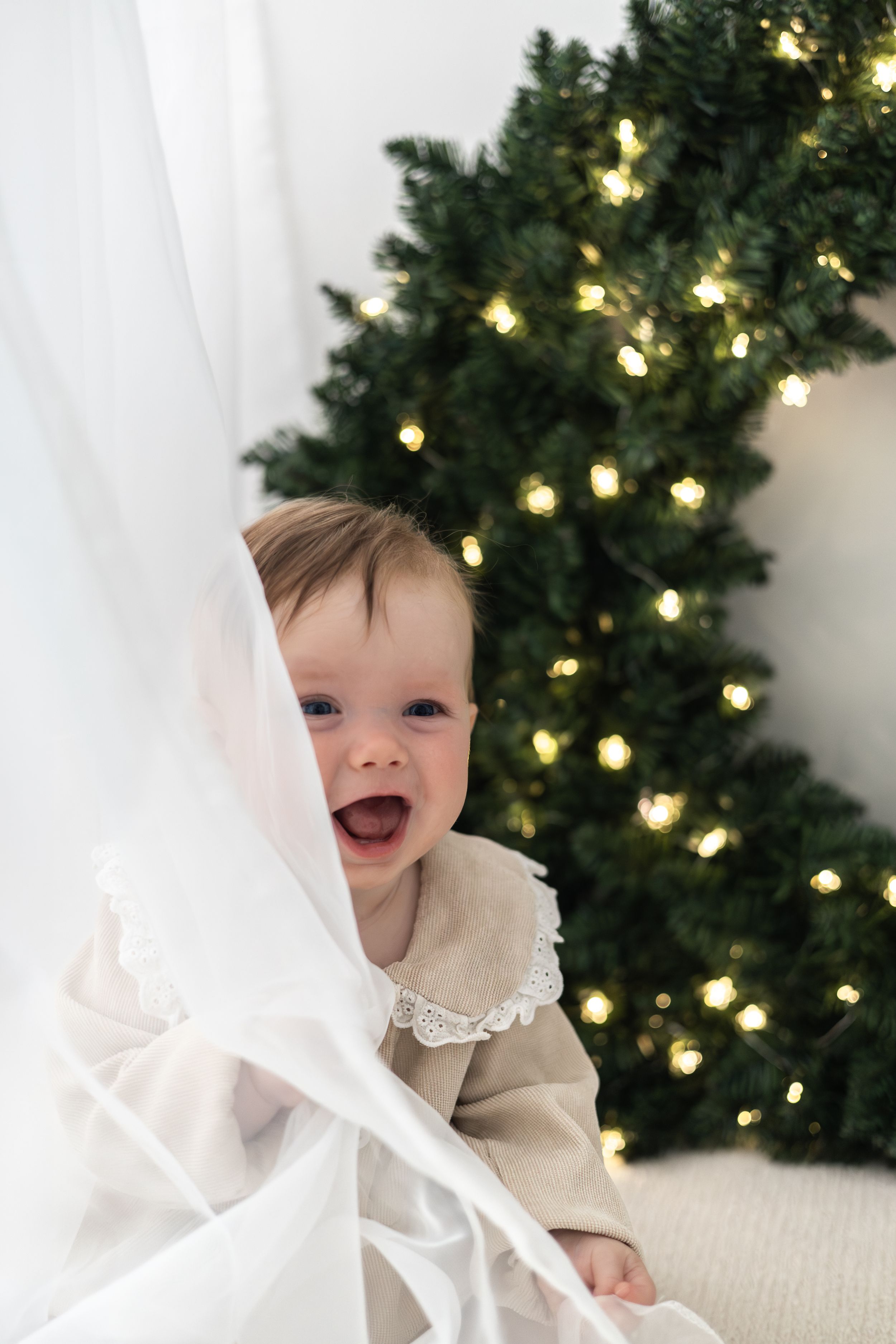 Joyful baby peeking through white curtain in front of Christmas wreath, Christmas mini Photoshoot Nottingham