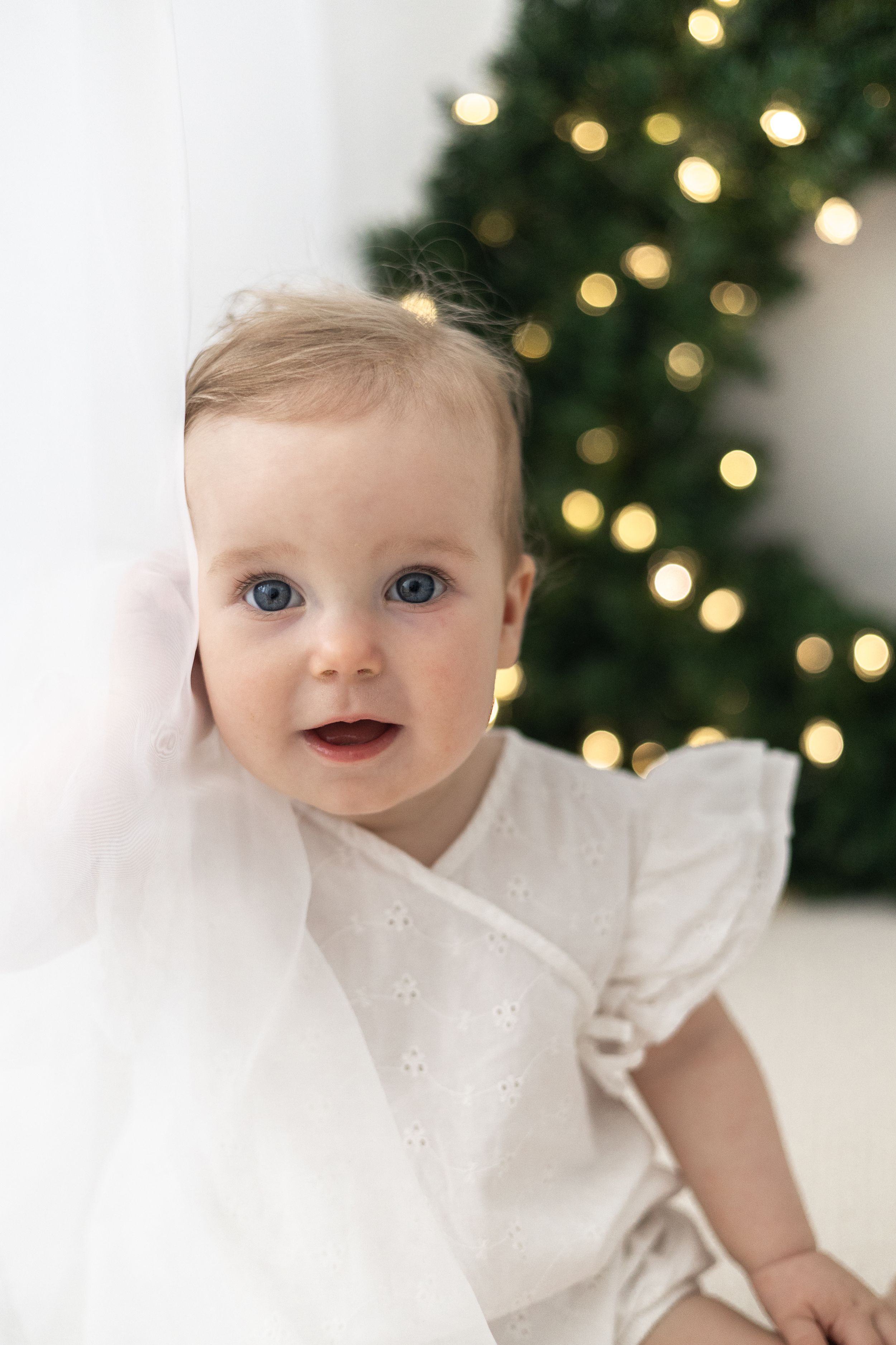 Smiling baby with blue eyes in white dress near lit Christmas wreath. Christmas mini Photoshoot Nottingham