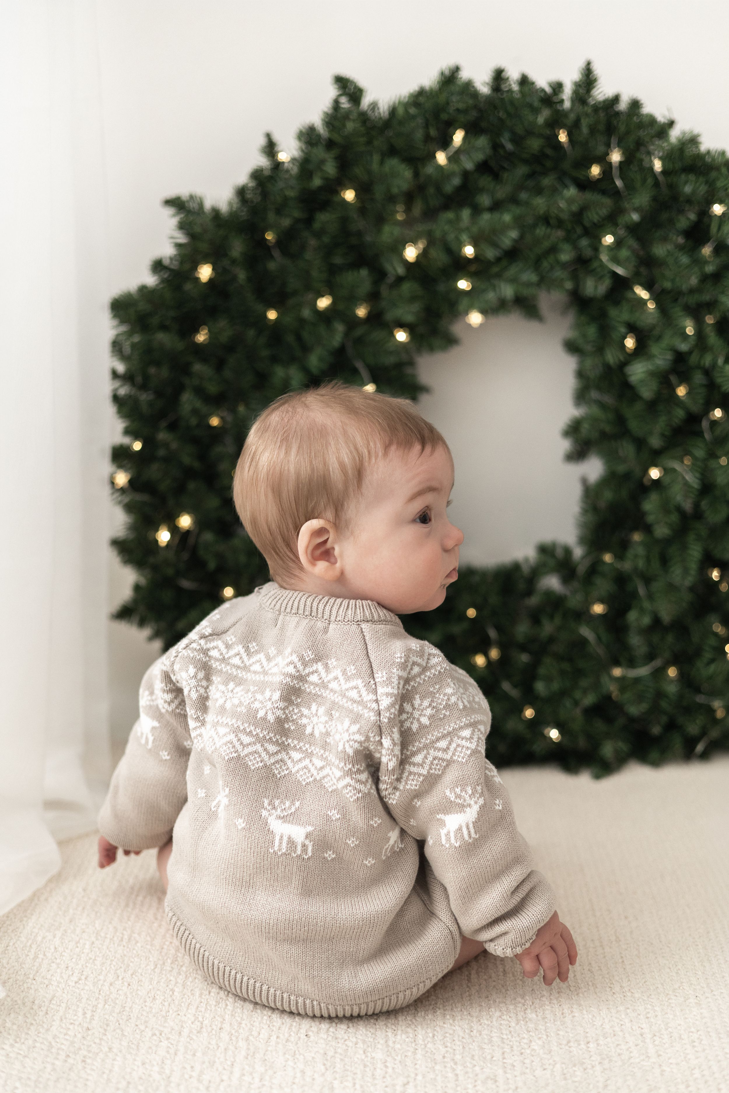 Baby in Nordic jumper sitting in front of decorated Christmas wreath with lights Christmas mini Photoshoot Nottingham