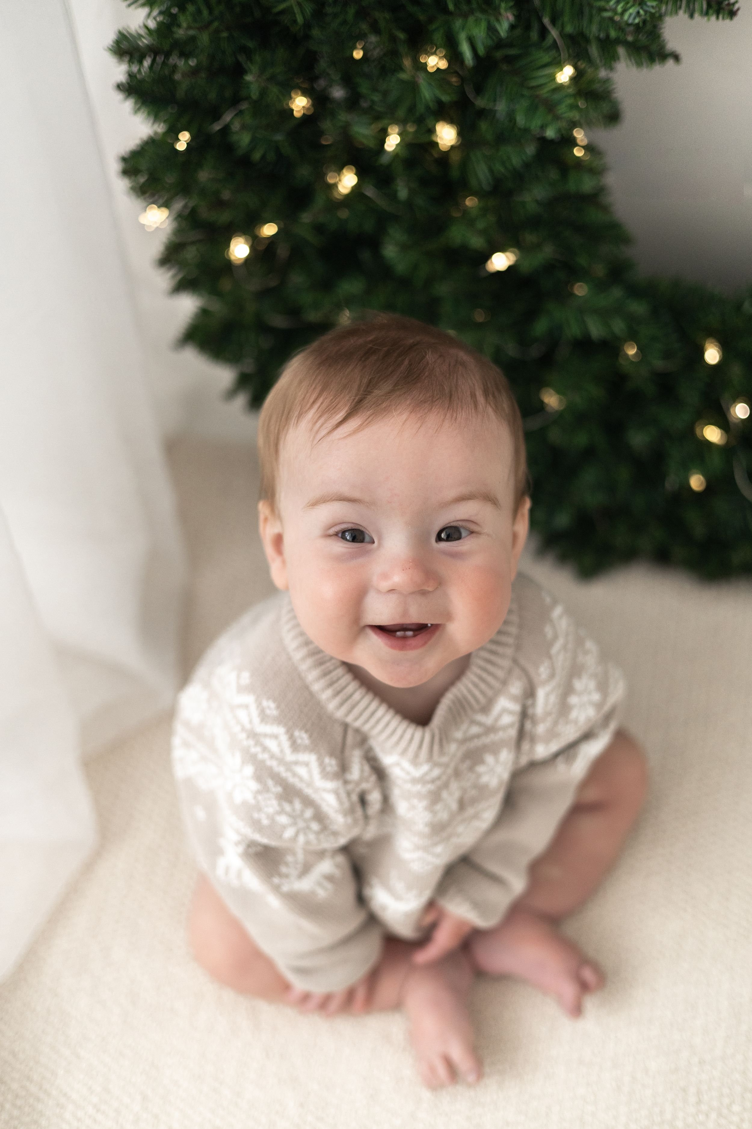 Smiling baby in a Nordic knit jumper sitting in front of a lit Christmas wreath on a white blanket Christmas mini Photoshoot Nottingham