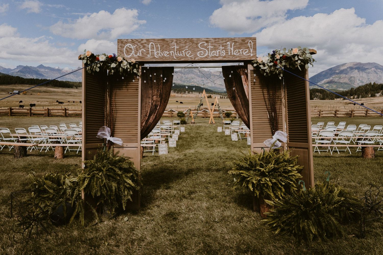 Outdoor wedding ceremony setup with wooden arch, floral decor, white chairs, and mountain backdrop on a sunny day.