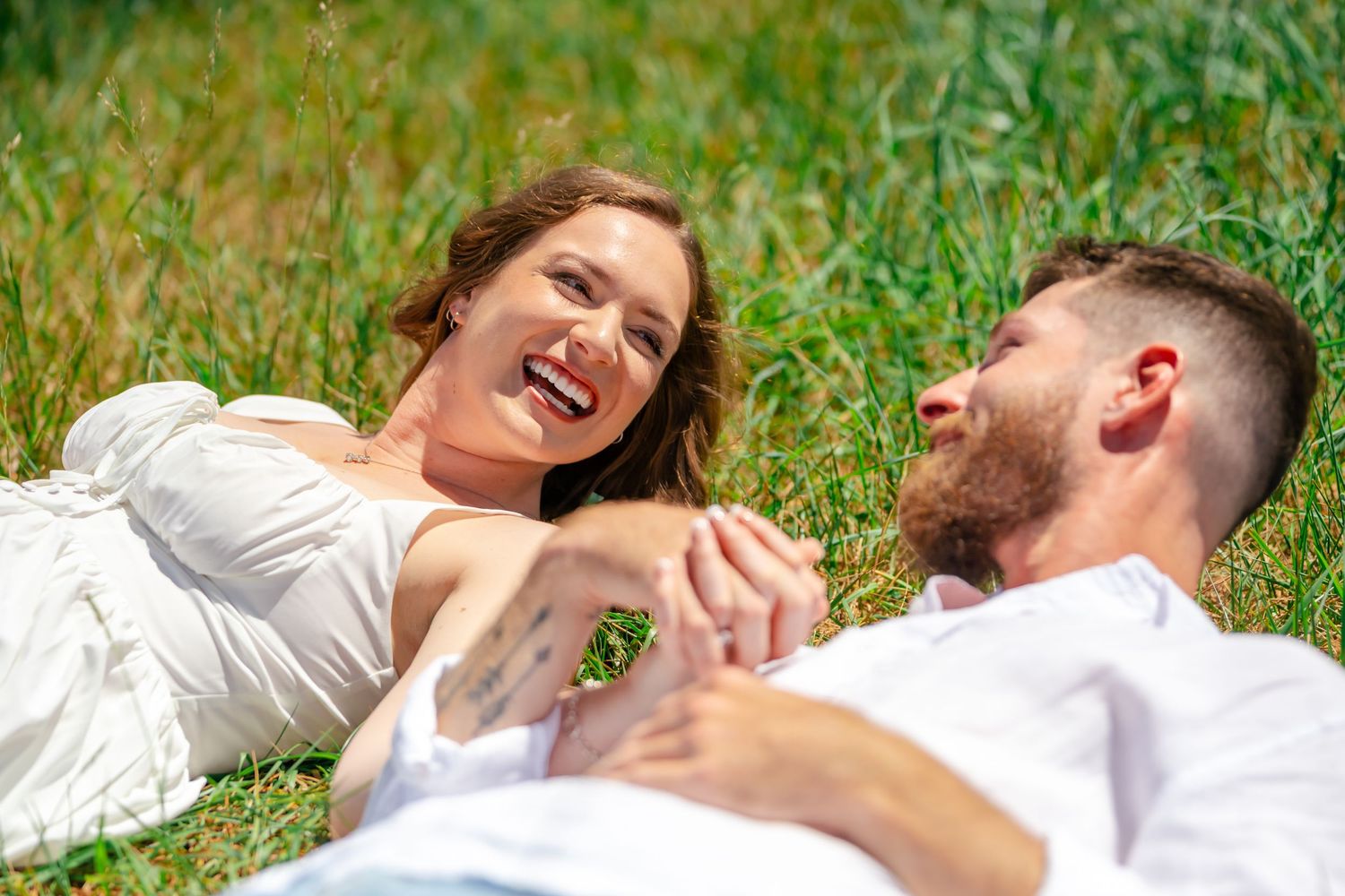 A happy couple lying in green grass, holding hands and smiling at each other on a sunny day.