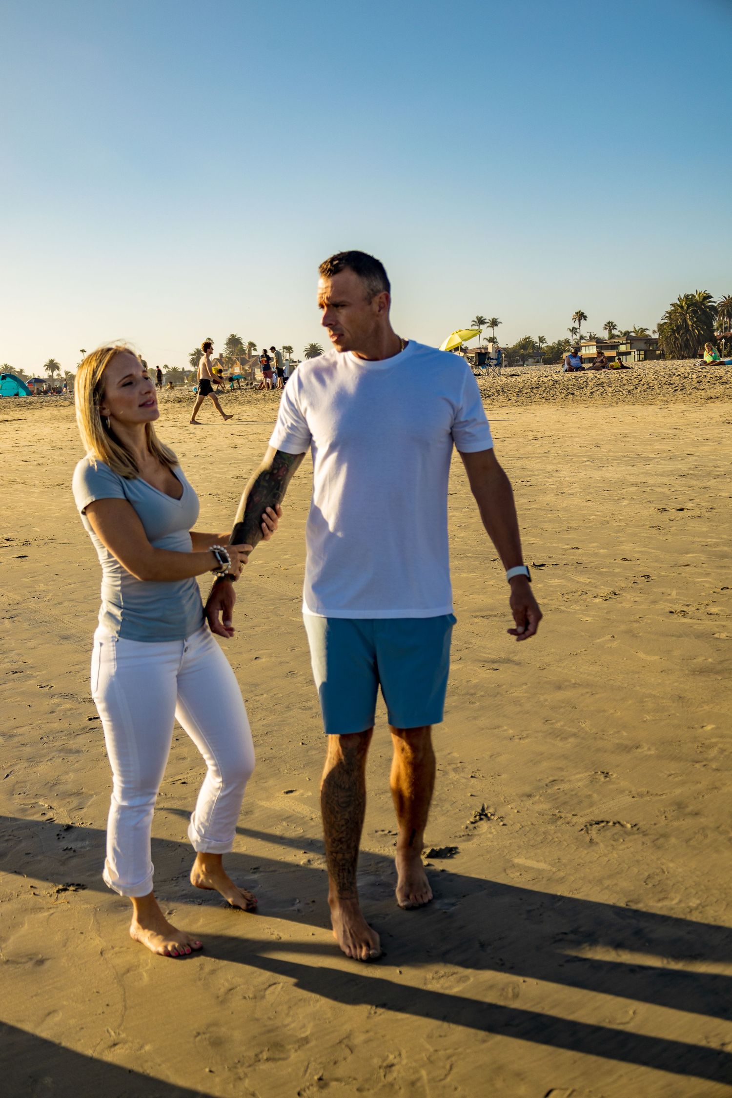 Couple holding hands and walking barefoot on a sunny beach, man in white tee and blue shorts, woman in light blue top and white jeans.
