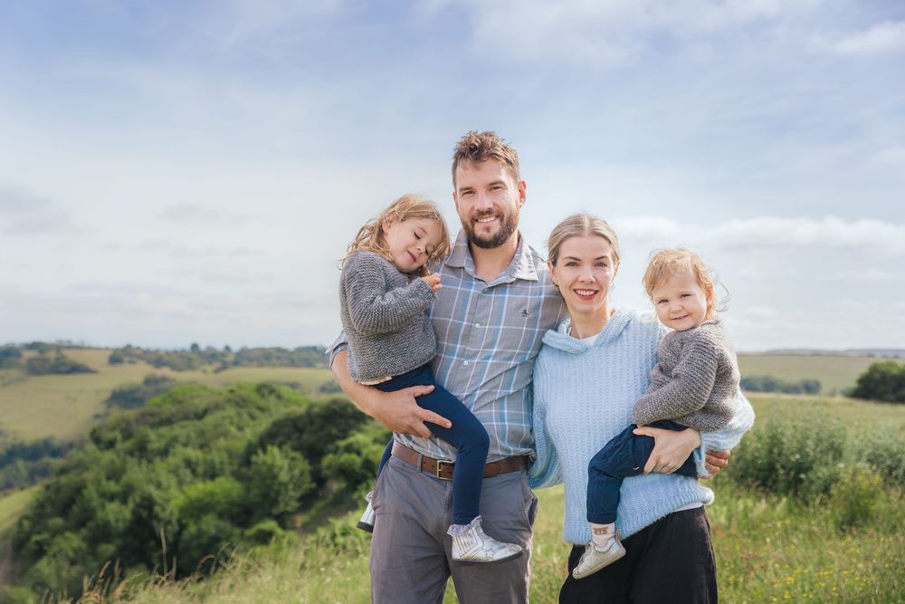 Happy family of four posing outdoors on a green hillside, parents holding two young children on a cloudy day.