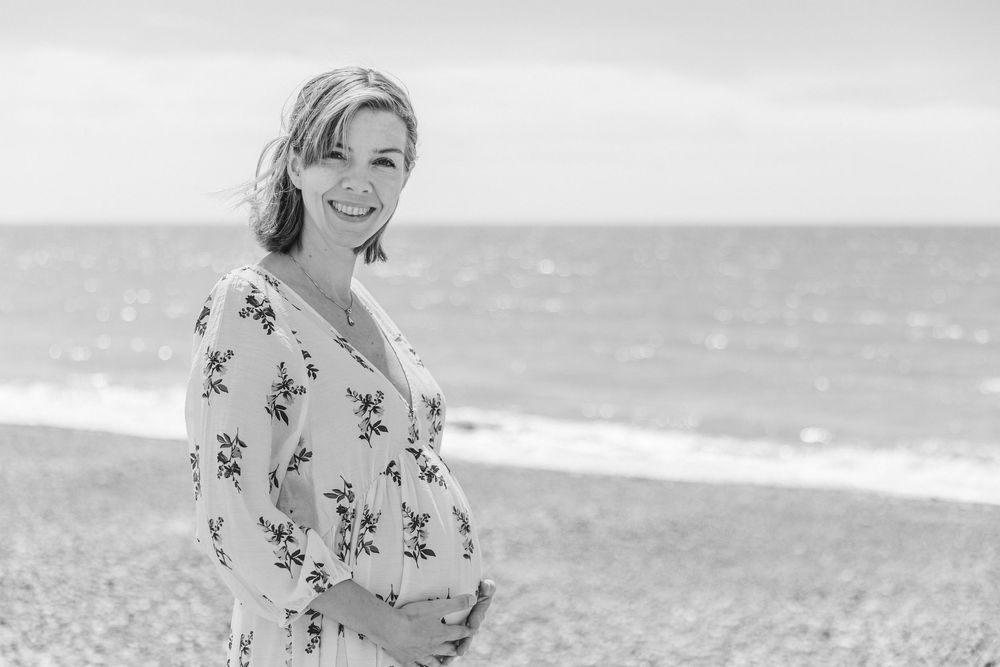 Smiling pregnant woman in floral dress cradling baby bump on a pebble beach in black and white.