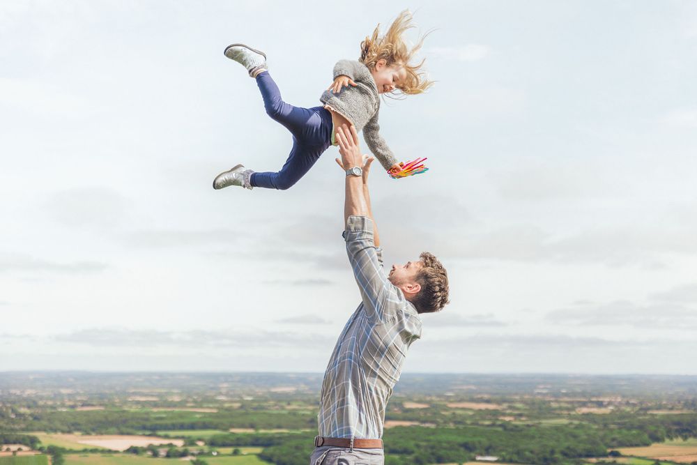Man lifting woman high in the air outdoors with scenic countryside landscape in the background.