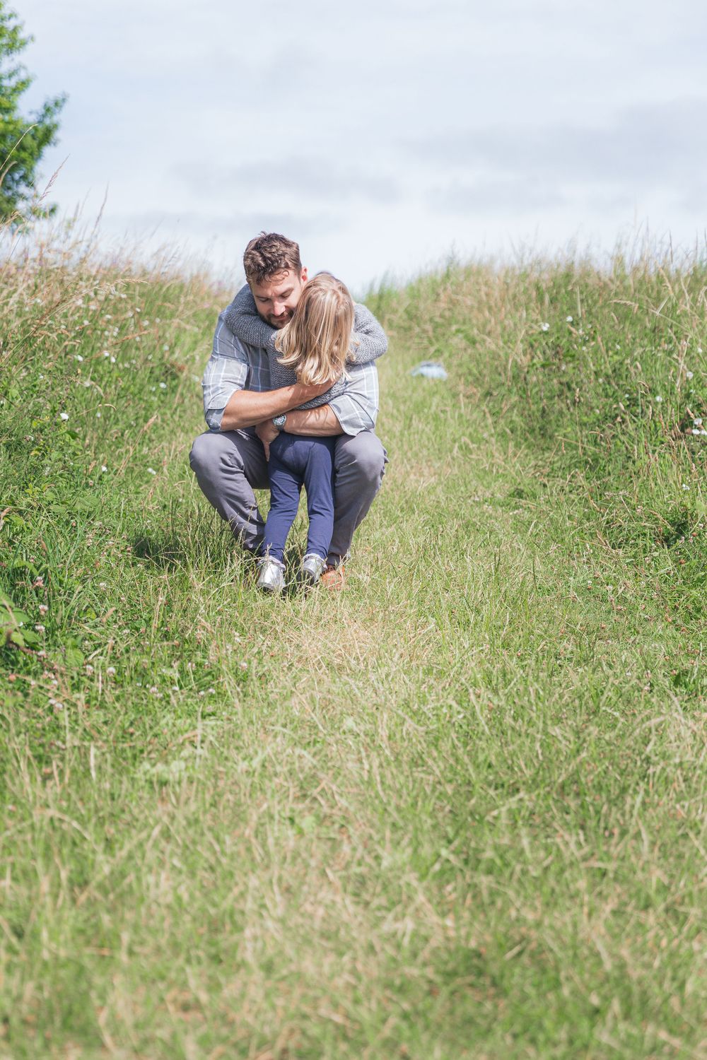 A man crouching in a green meadow, warmly embracing a young child on a cloudy day.