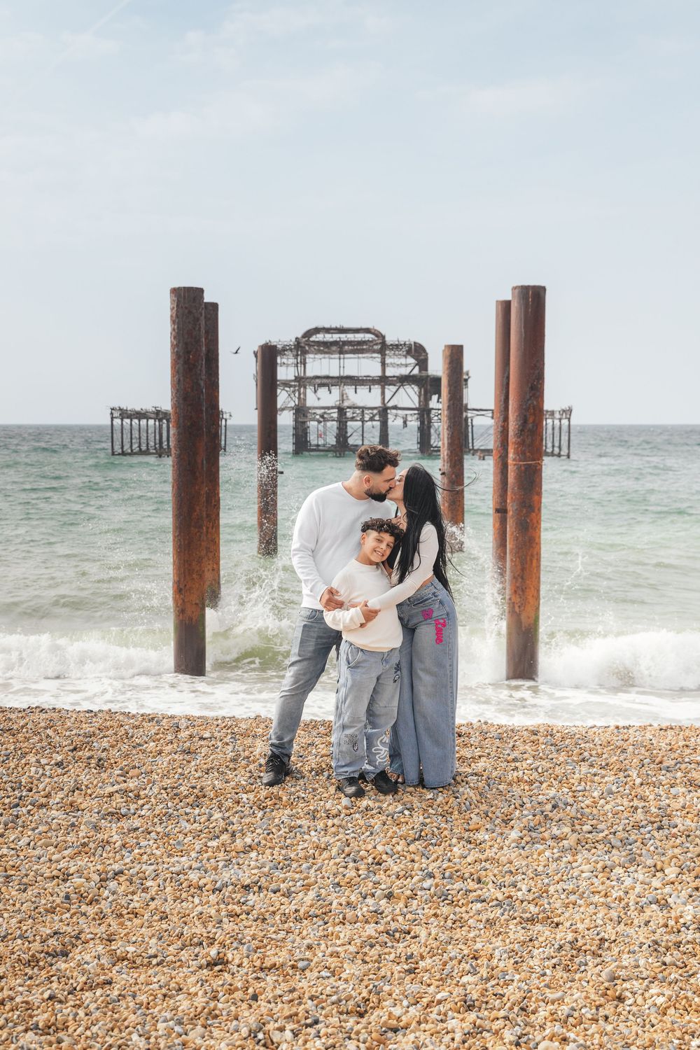 Family of three embracing on Brighton pebble beach with the iconic ruined West Pier structure behind them.