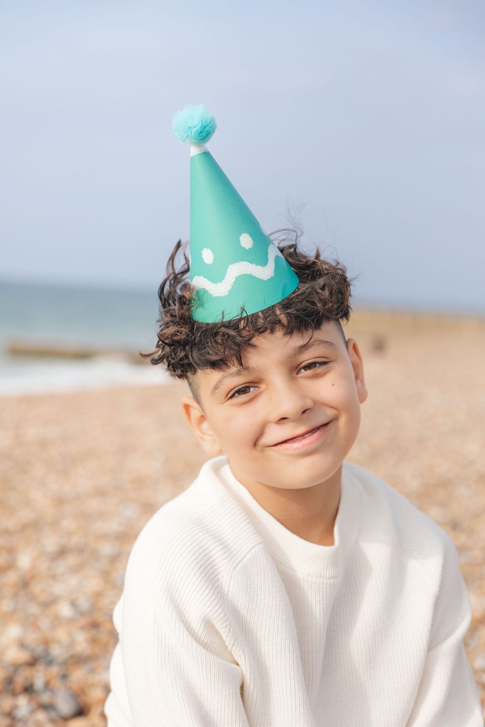 Smiling boy wearing a teal birthday party hat on a sunny beach, dressed in a white sweater.