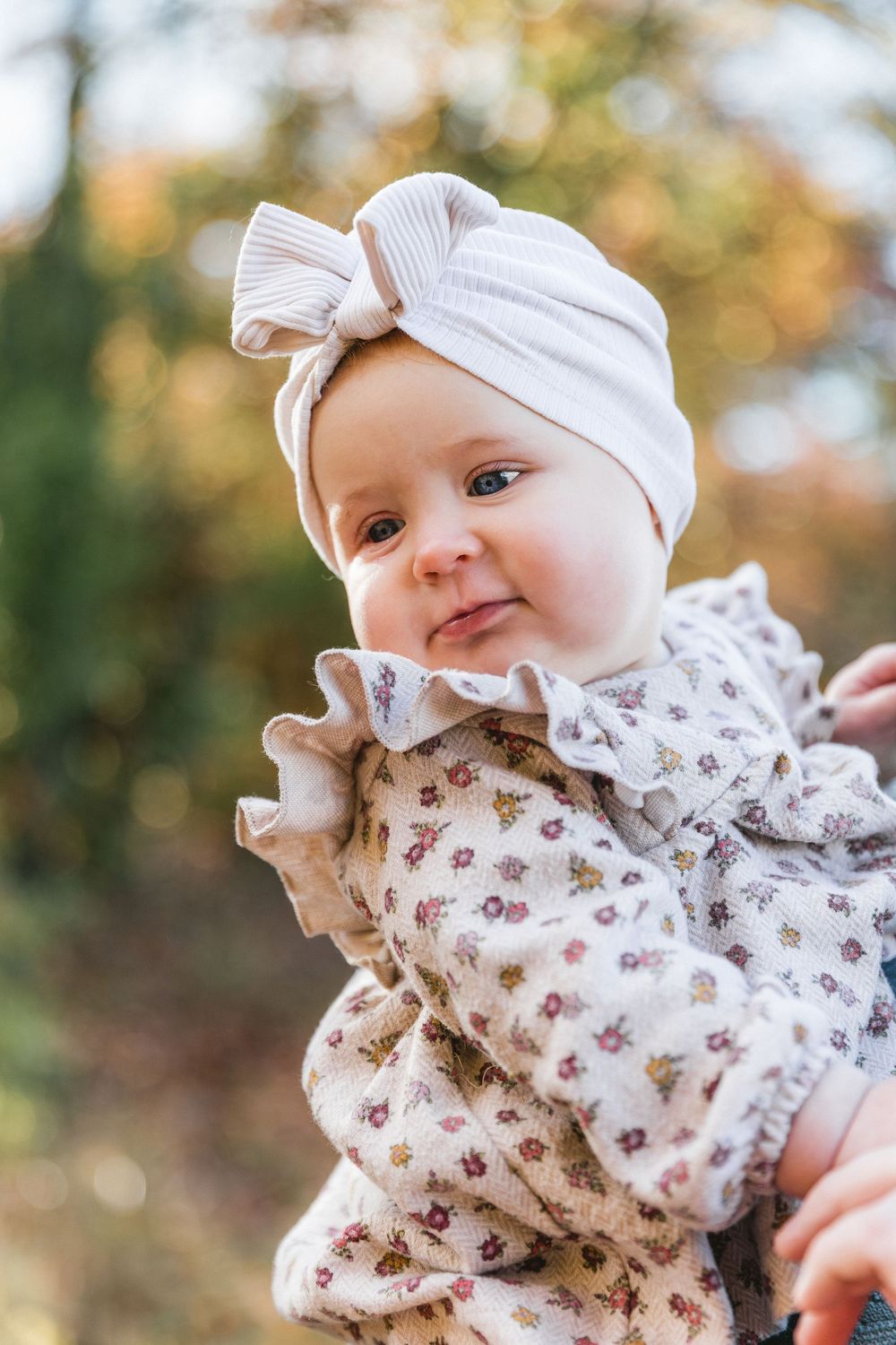 Adorable baby girl in floral outfit and white bow turban hat smiling outdoors in soft sunlight.