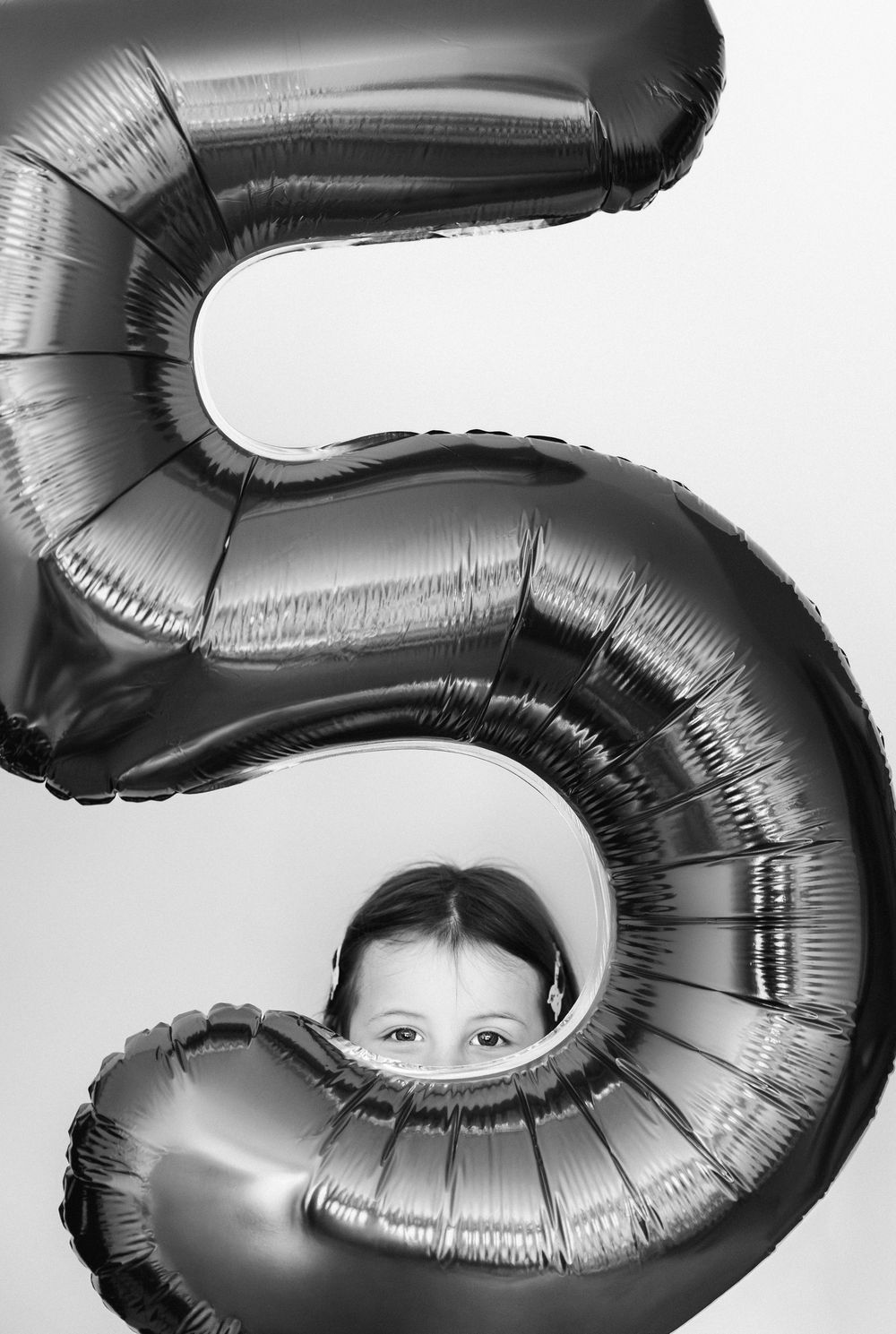 Child playfully hiding behind large silver number 5 balloon in black and white birthday photo.