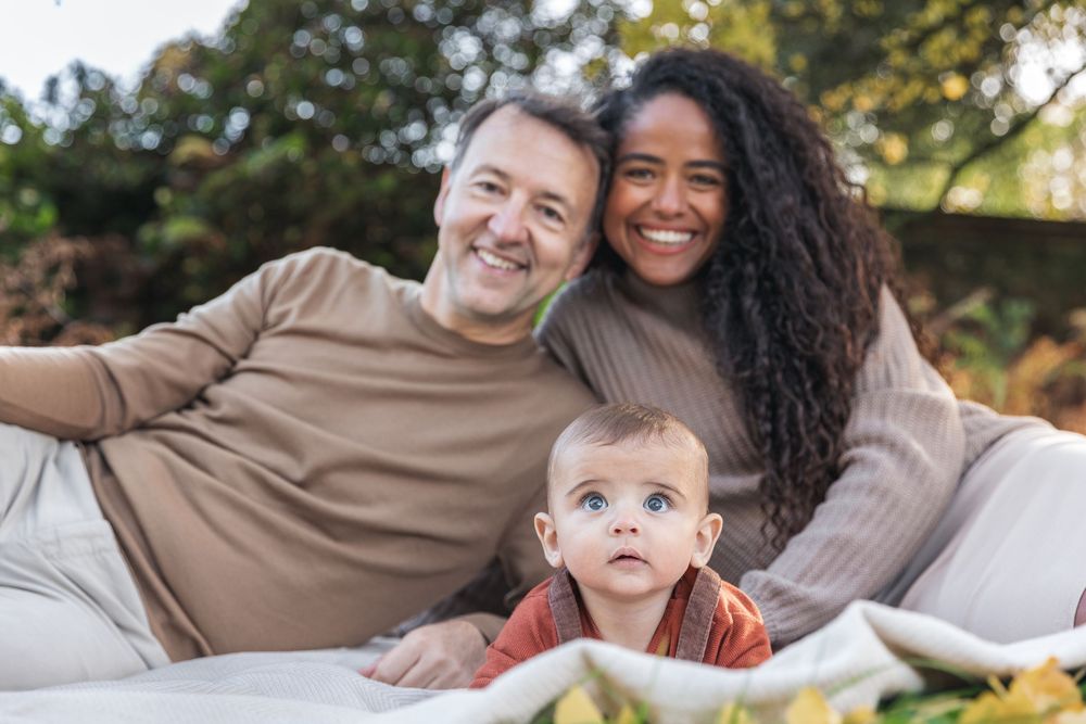 Smiling interracial couple lying outdoors with curious blue-eyed baby between them in autumn setting.