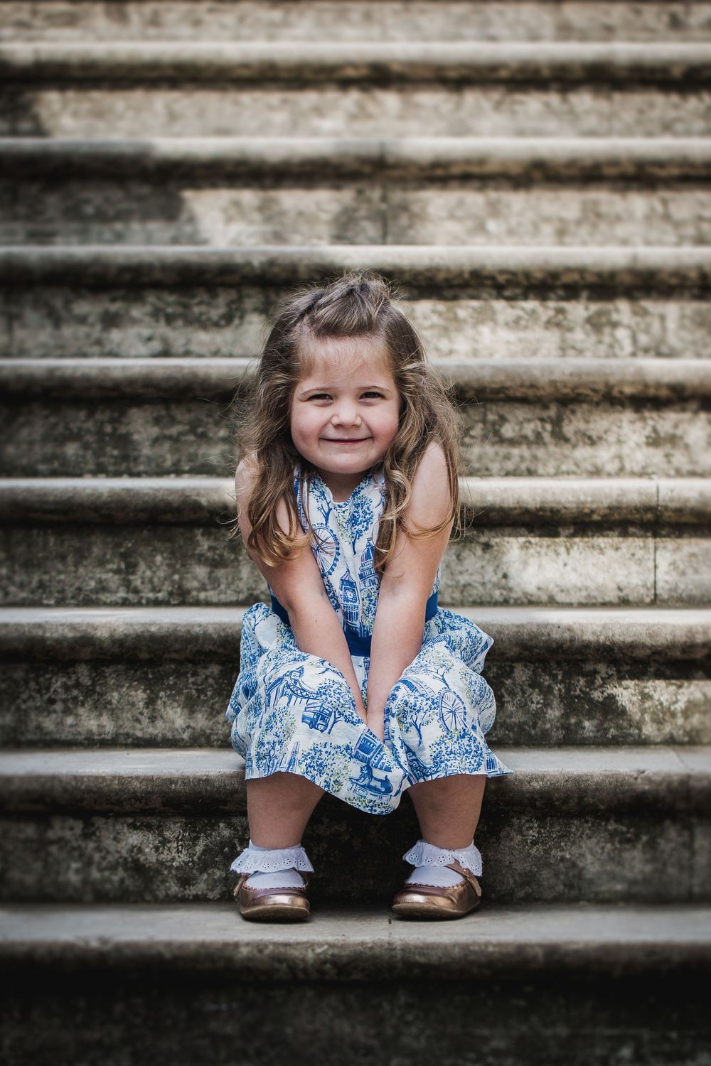 Cute little girl in a blue floral dress sitting on stone steps smiling sweetly.