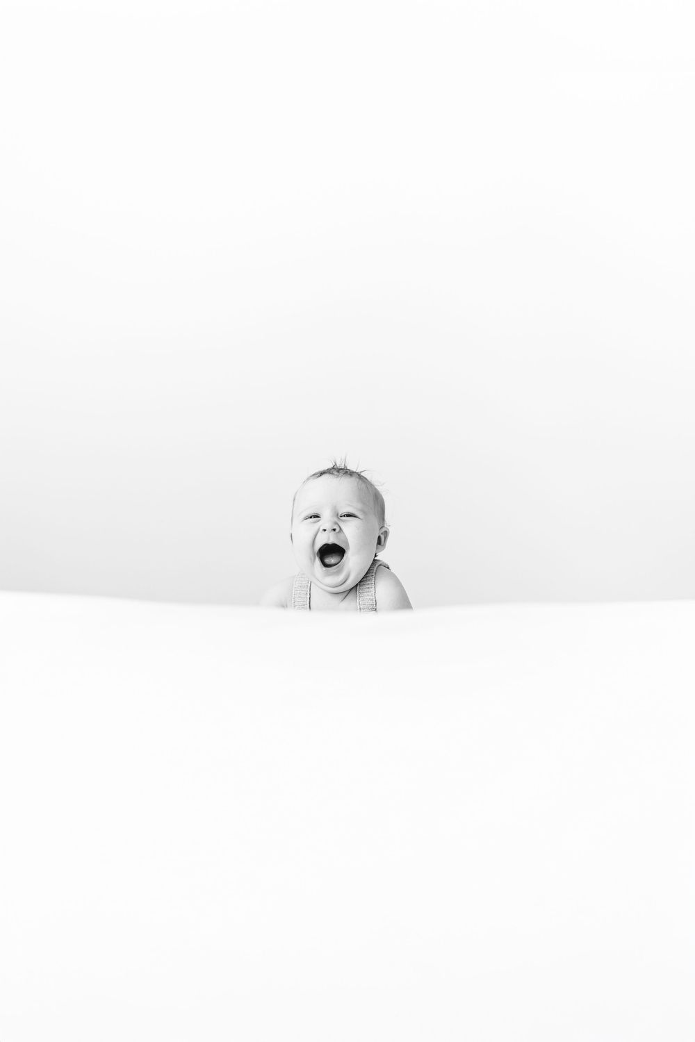 Black and white minimalist photo of yawning newborn baby peeking above white surface.