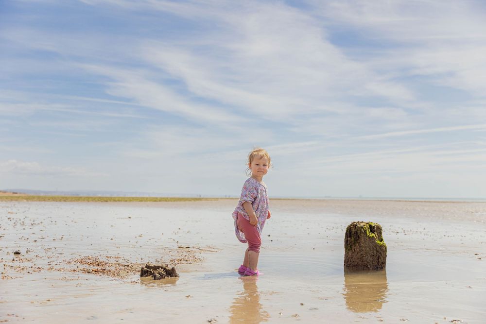 Young child in pink outfit and rain boots standing on a shallow sandy beach under a blue sky.