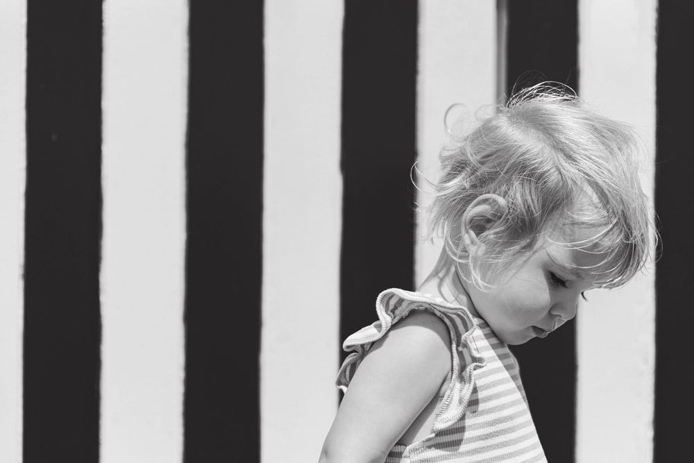 Black and white portrait of toddler girl with windswept hair against bold striped background.