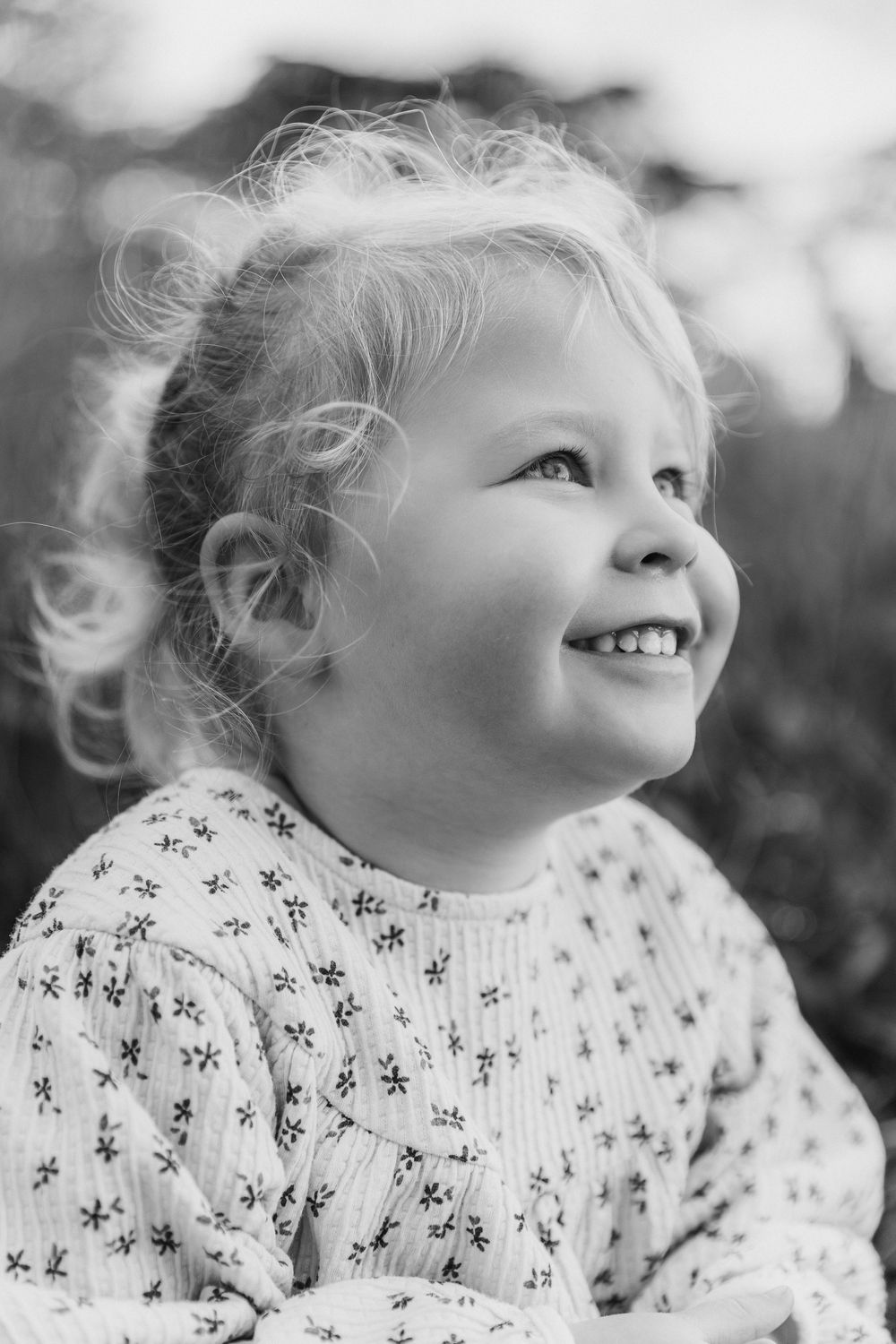 Black and white portrait of smiling blonde toddler girl in floral dress looking up joyfully.