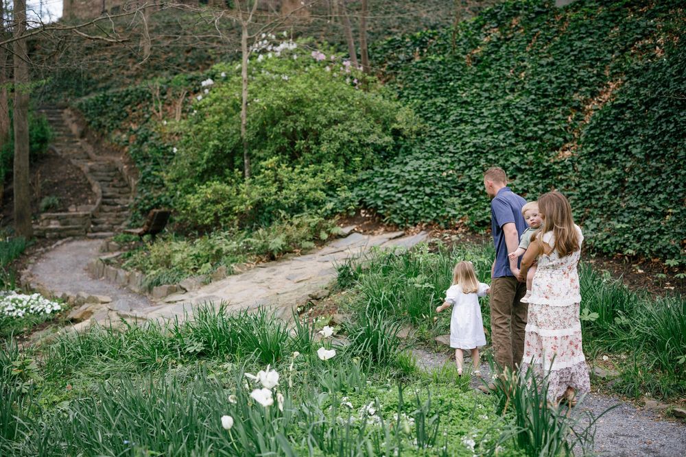 Family of three standing on a garden path surrounded by lush greenery and white flowers in bloom.