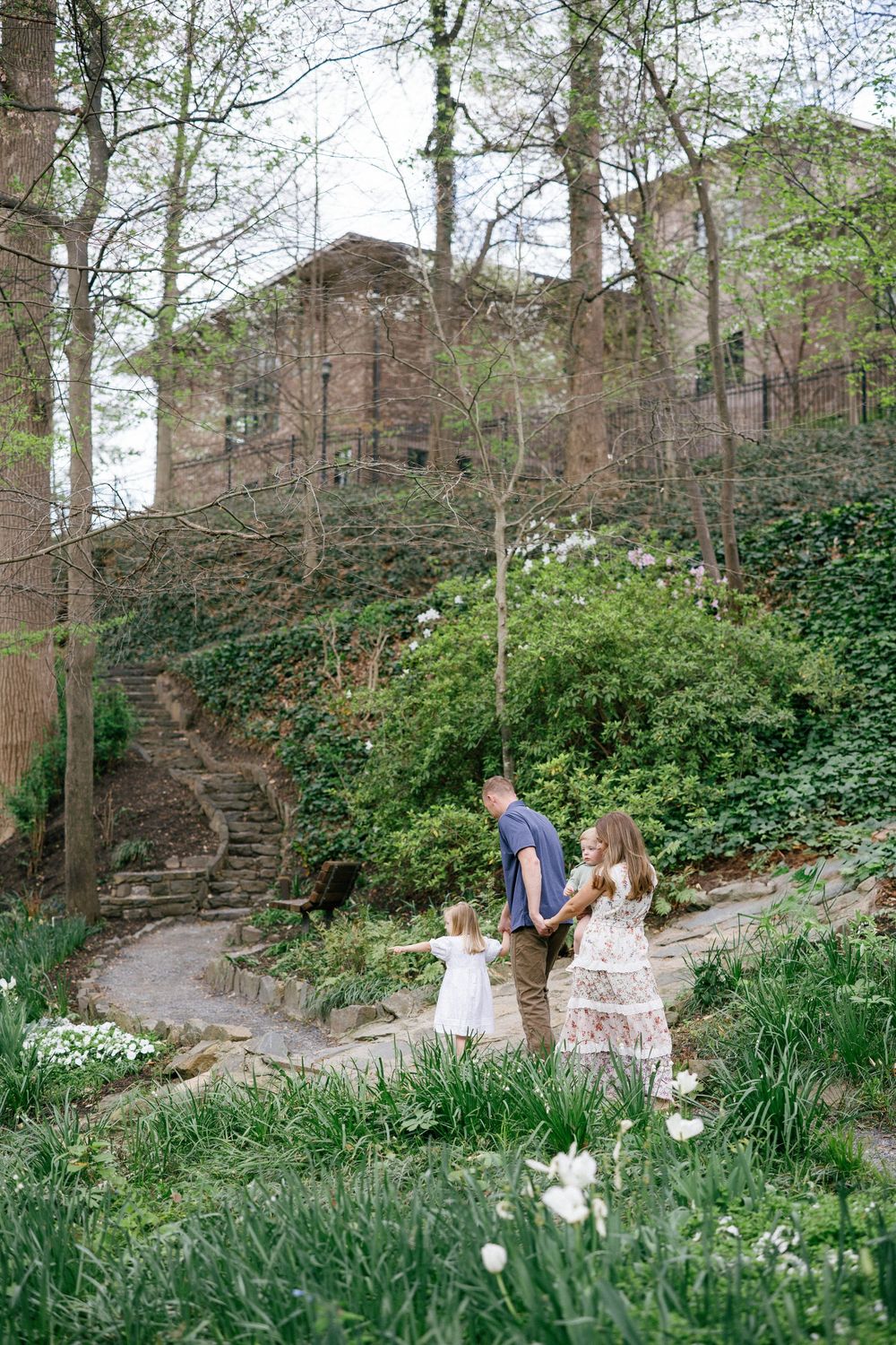 Family of three walking on a stone path in a lush green garden with ivy-covered walls and spring blooms.