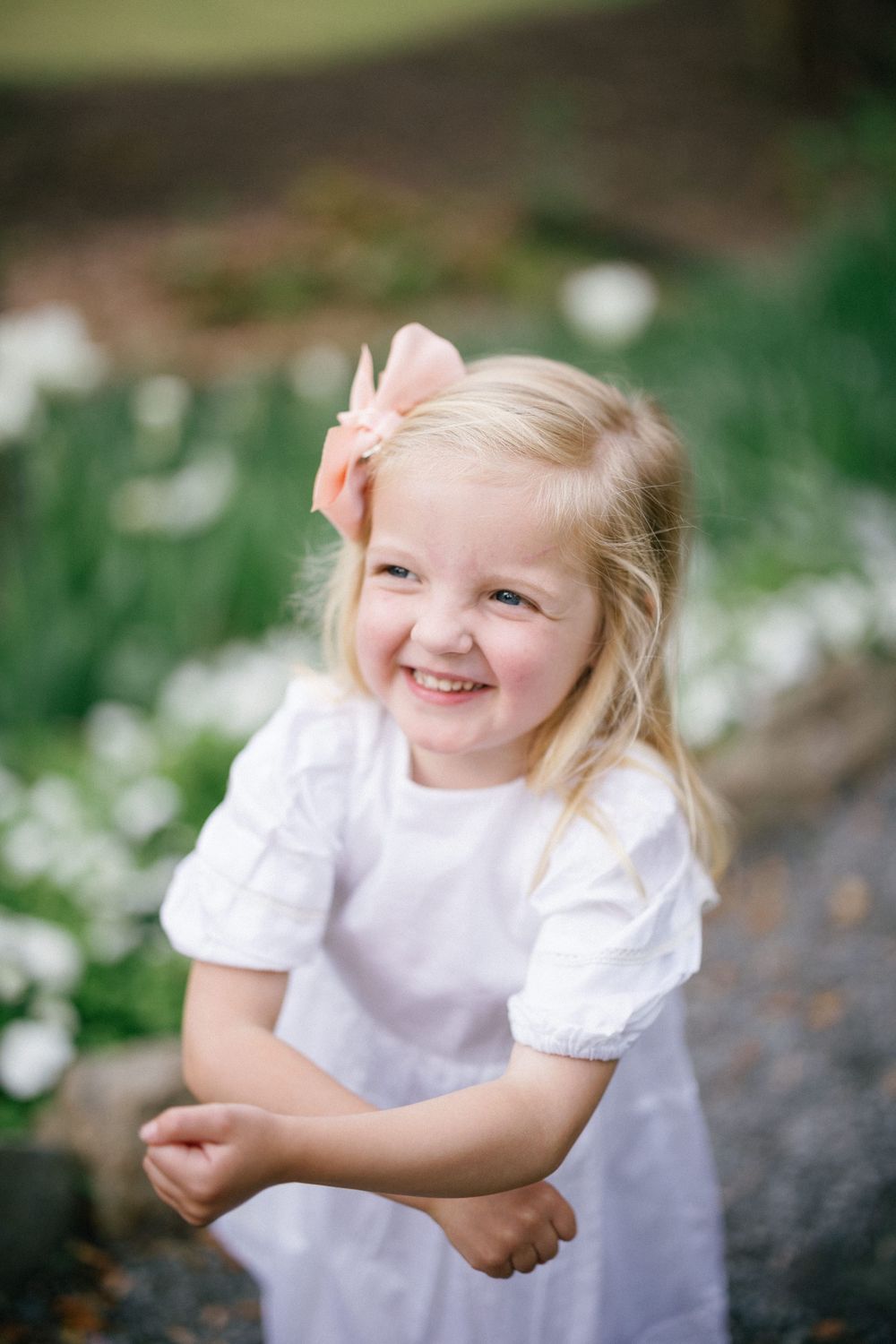 Smiling blonde toddler girl in white dress with pink bow, posing outdoors near white flowers in a garden.