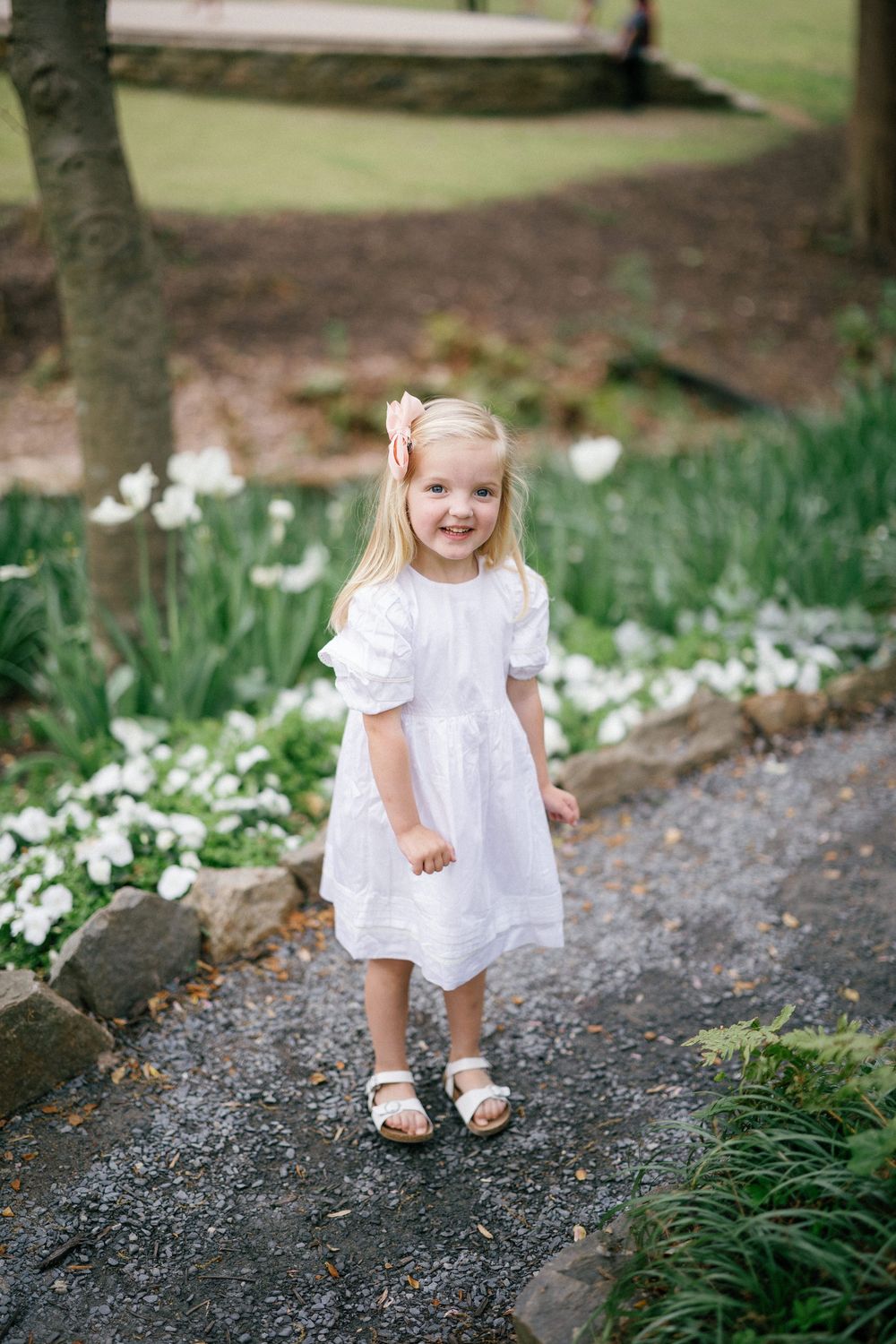 A smiling little girl in a white dress and sandals walks along a garden path surrounded by white flowers.