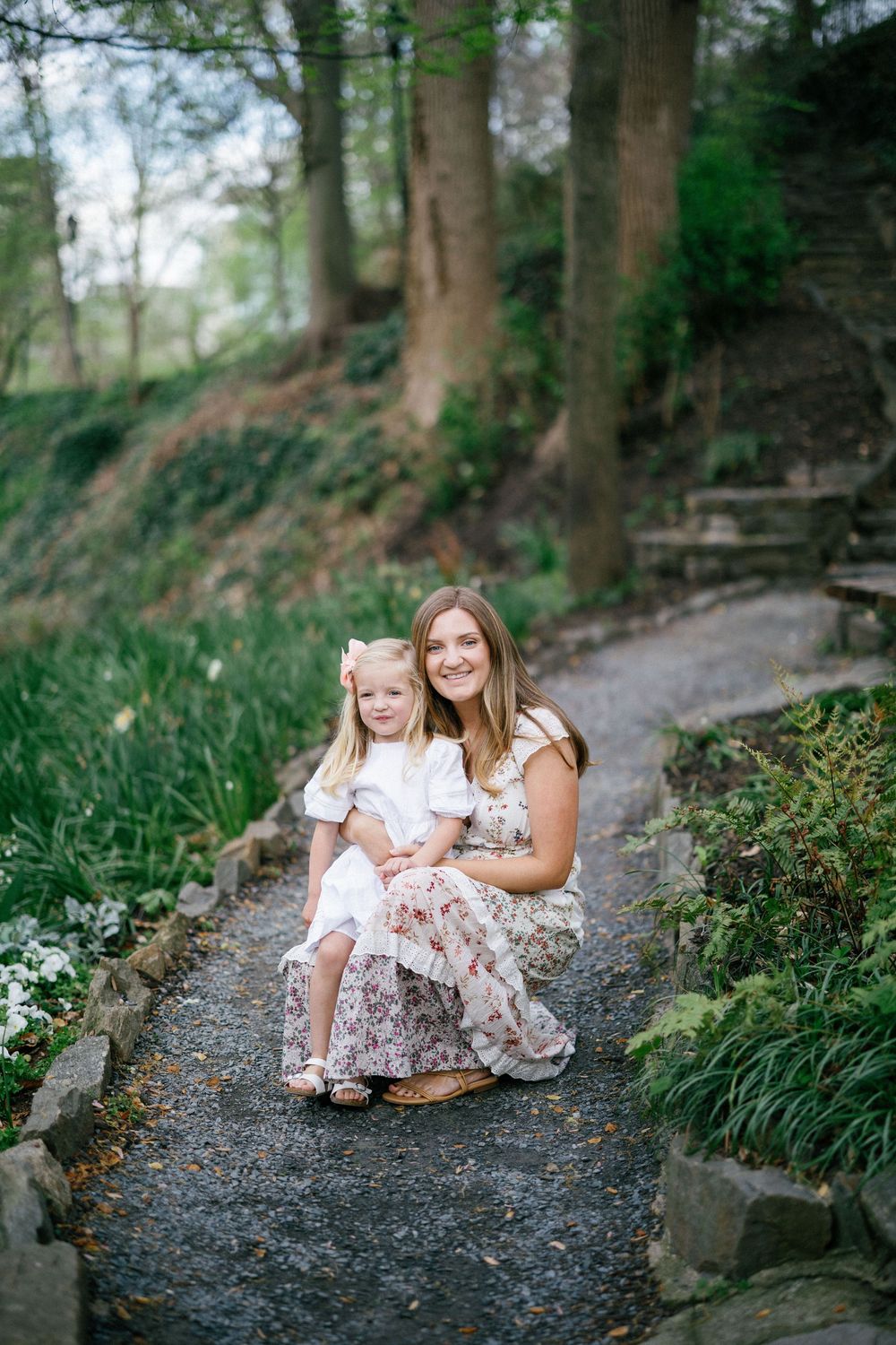A smiling mother crouches on a garden path hugging her young blonde daughter in a white dress among lush greenery.