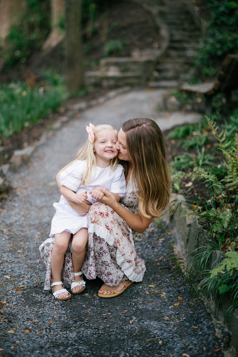 A mother crouching on a garden path, kissing her smiling blonde toddler daughter in white dress.