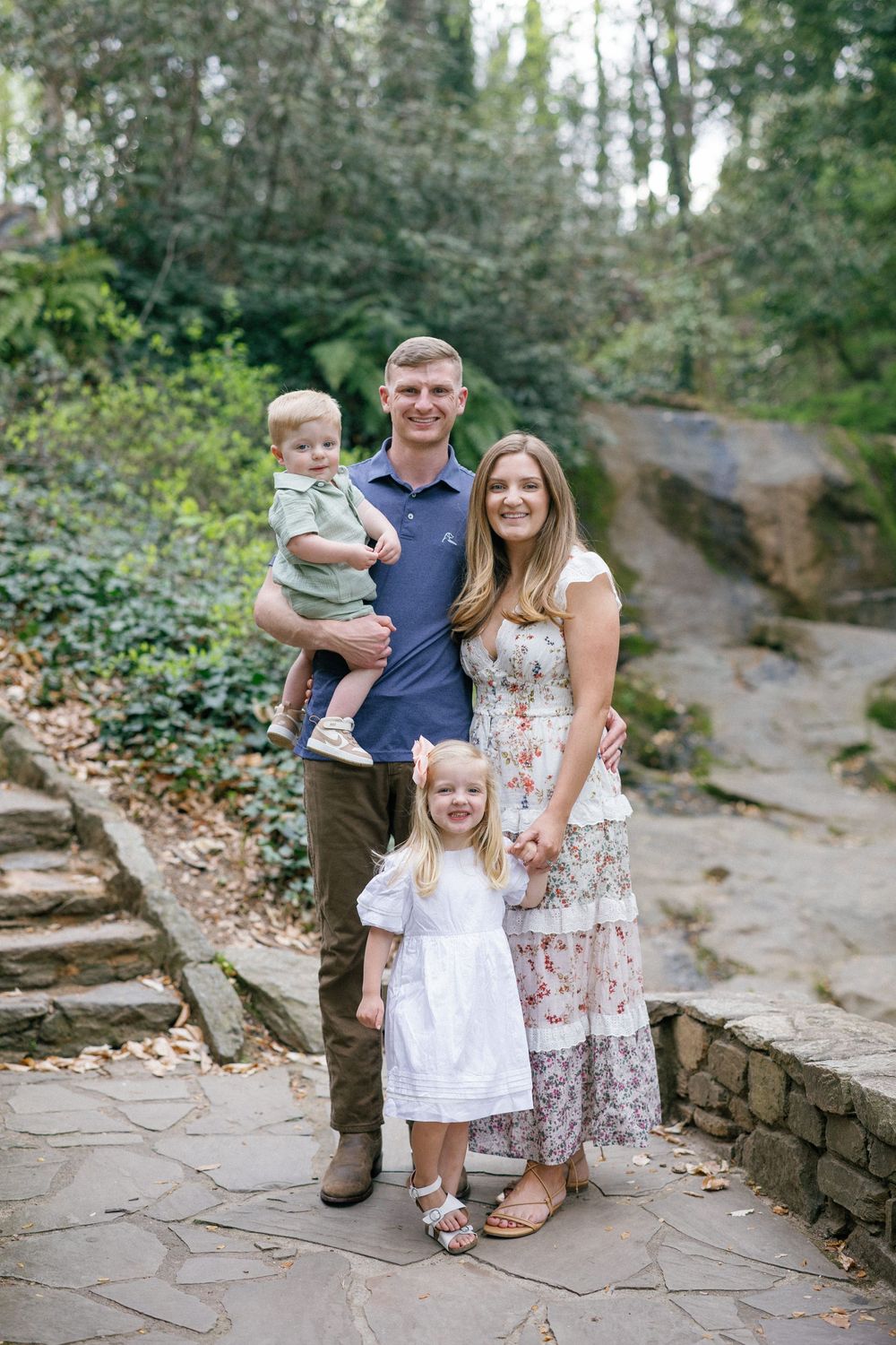 A happy family of four poses on stone steps surrounded by lush greenery, parents holding young children.