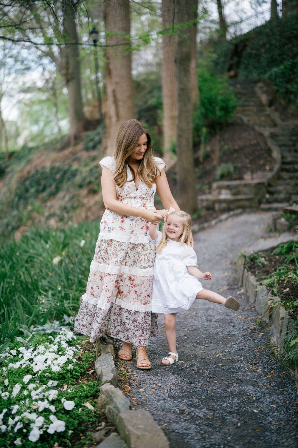 Mother in floral dress holds hands with young daughter in white dress on a garden path surrounded by spring blooms.
