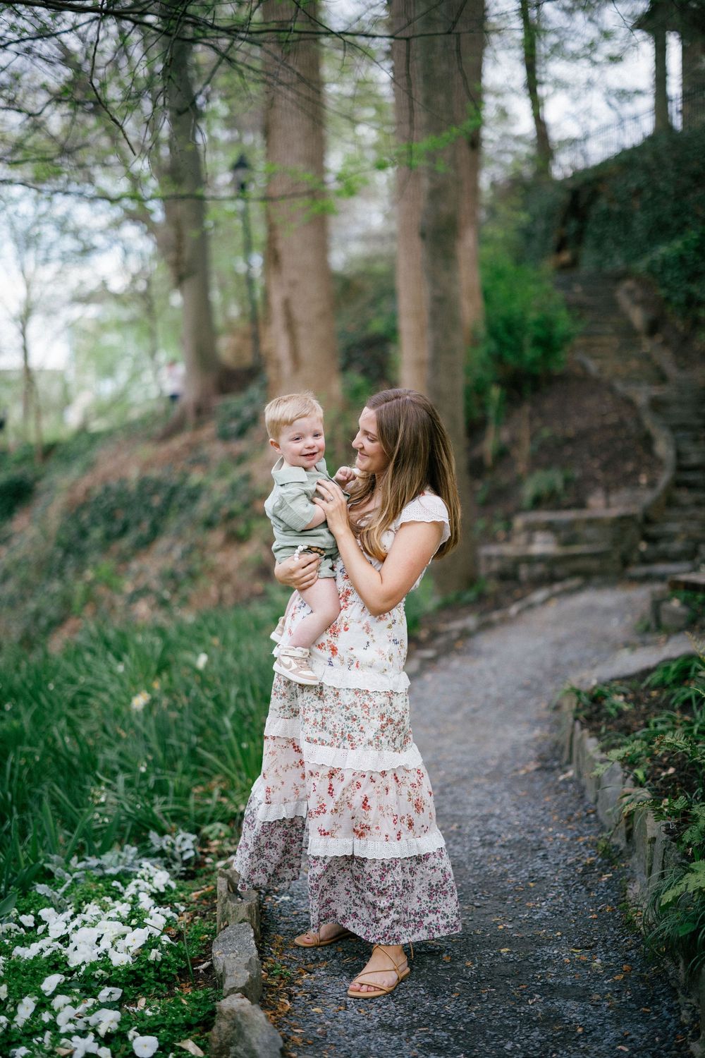 A mother in a floral maxi dress holds her toddler on a garden path surrounded by lush greenery and white flowers.