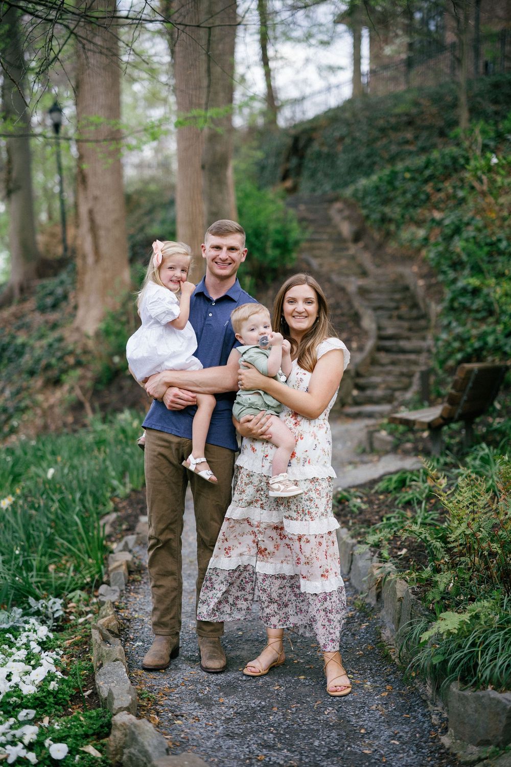 Young family of four posing on a garden path surrounded by lush greenery and stone steps in spring.