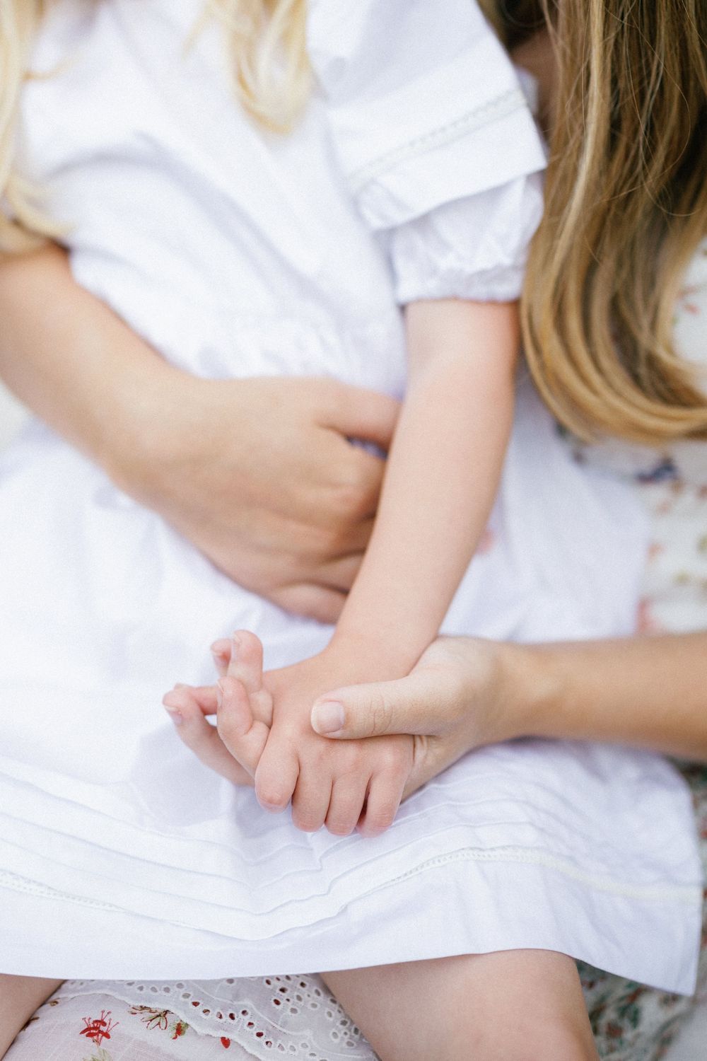 A mother in a white dress gently holds her young daughter's hands, both wearing white dresses in a tender moment.