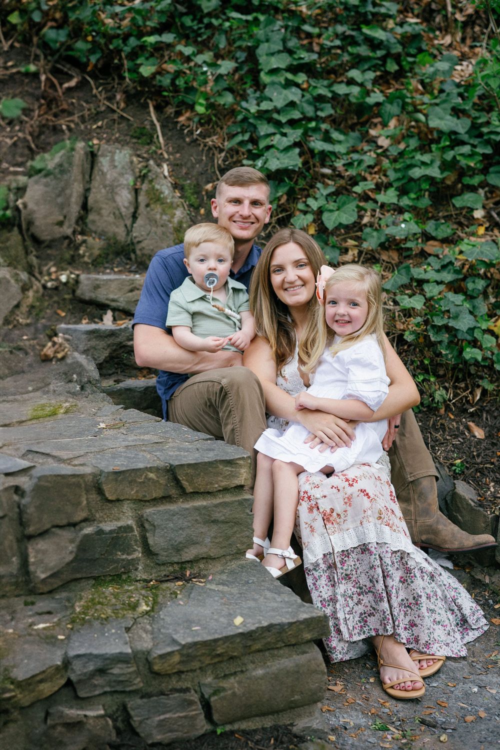 A smiling family of four poses on stone steps surrounded by lush greenery, parents holding two young children.