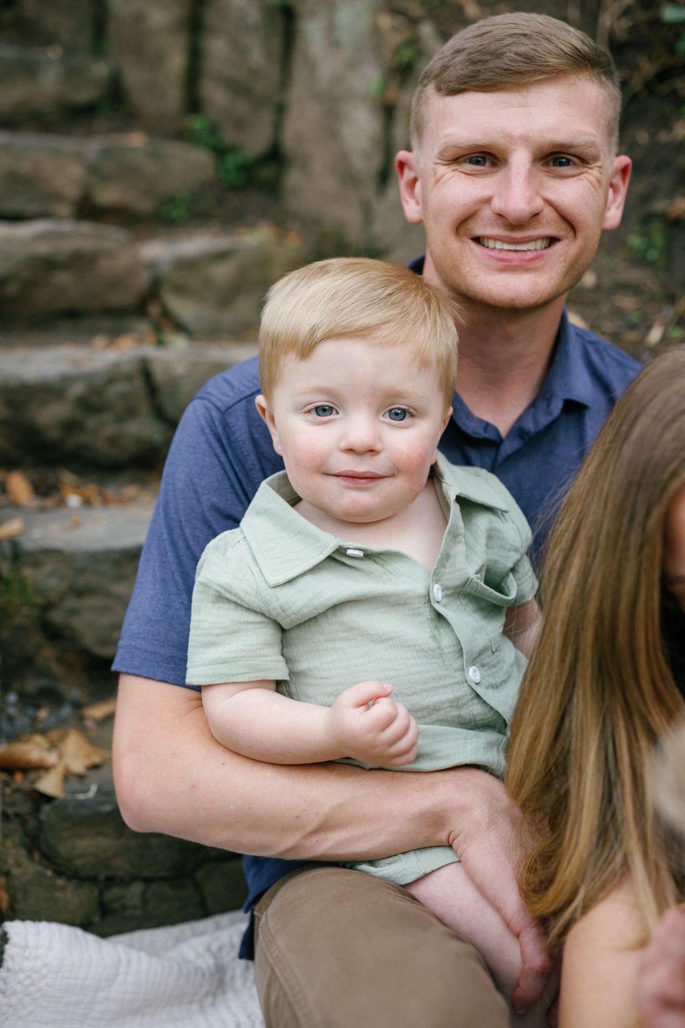 Smiling man holding a blonde toddler boy with blue eyes outdoors near stone steps.
