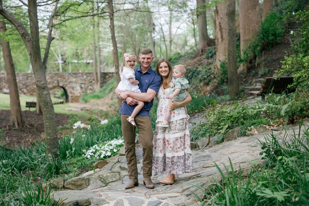 Happy family of four posing on a stone path in a lush green garden with a rustic bridge backdrop.